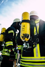 A group of firefighters wearing helmets and protective gear, including bright yellow oxygen tanks strapped to their backs. The reflective stripes on their suits are visible, and they seem to be preparing for or engaged in an operation outdoors under a clear sky.