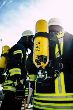 A group of firefighters wearing helmets and protective gear, including bright yellow oxygen tanks strapped to their backs. The reflective stripes on their suits are visible, and they seem to be preparing for or engaged in an operation outdoors under a clear sky.
