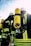 A group of firefighters wearing helmets and protective gear, including bright yellow oxygen tanks strapped to their backs. The reflective stripes on their suits are visible, and they seem to be preparing for or engaged in an operation outdoors under a clear sky.