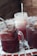 Three mason jars filled with a dark reddish drink sit on a mosaic table. The drinks appear to be partially frozen or slushy, and each jar has a metal straw. The jars are arranged close to each other, with condensation visible on the glass surfaces.