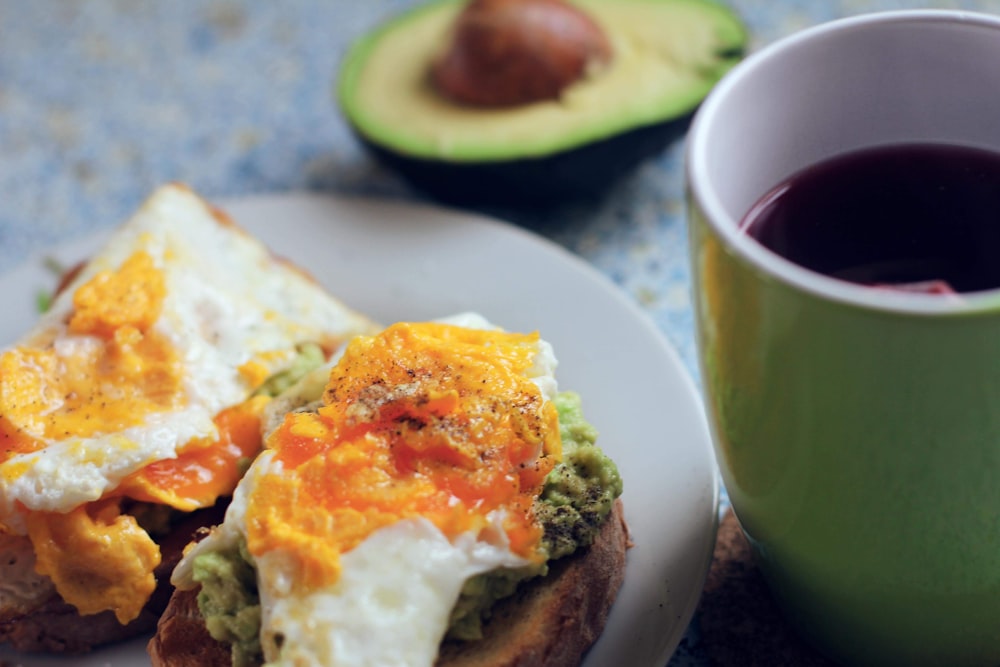 plate of breakfast sandwich beside avocado and hot beverage