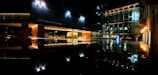 A nighttime scene of a modern cultural center with large glass windows illuminated by exterior lighting. The reflection of the building and lights can be seen on a glossy surface below, enhancing the architectural design.