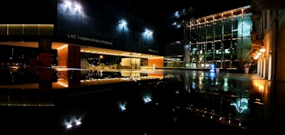 A nighttime scene of a modern cultural center with large glass windows illuminated by exterior lighting. The reflection of the building and lights can be seen on a glossy surface below, enhancing the architectural design.