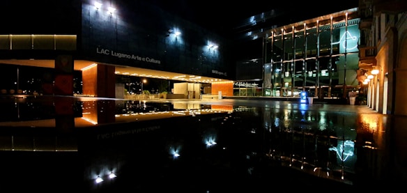 A nighttime scene of a modern cultural center with large glass windows illuminated by exterior lighting. The reflection of the building and lights can be seen on a glossy surface below, enhancing the architectural design.