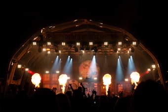 A nighttime concert stage with bright spotlights and pyrotechnic flames shooting up from the ground. The stage is adorned with lights and a backdrop displaying artwork. A crowd of silhouetted figures raises their hands in the foreground.