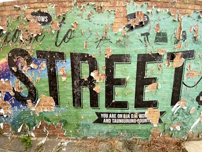 A weathered and peeling mural on a brick wall with the word 'STREET' prominently displayed in large black letters. The mural has a distressed appearance with layers of paint chipping away, revealing a mix of colors underneath. The background features a faded green and blue gradient with elements of urban art such as symbols and stylized text. Grass is visible at the bottom of the wall.
