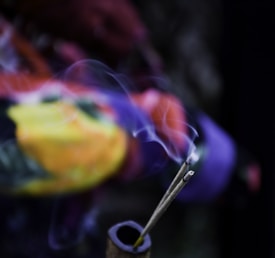 Two incense sticks emit delicate streams of smoke against a blurred, colorful background. The smoke swirls gracefully and creates a soft, ethereal atmosphere.