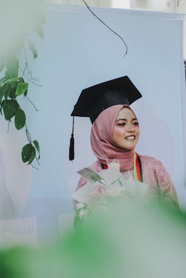 A young woman wearing a graduation cap and a hijab is holding a bouquet of flowers. She is smiling, and her attire is complemented by a decorative stole. The background features soft pastel colors, with greenery framing the scene.