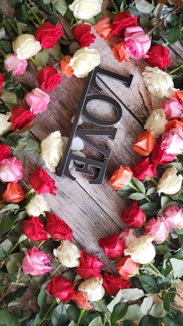 A decorative arrangement of multi-colored roses forming a circular pattern on a wooden surface, surrounding a metallic decorative object spelling out the word 'LOVE' in capital letters.