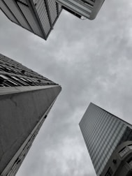 Three tall skyscrapers converge from different angles against an overcast sky, creating a sense of depth and urban architecture. The buildings feature a mixture of glass and concrete facades.