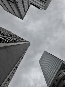 Three tall skyscrapers converge from different angles against an overcast sky, creating a sense of depth and urban architecture. The buildings feature a mixture of glass and concrete facades.