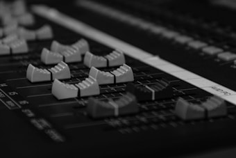 A close-up view of a professional audio mixing console with multiple sliders and knobs. The sliders are aligned in a row, each with textured grips for adjustment. The surface of the console is matte black with white markings indicating different channels.