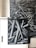 A black and white photograph featuring a complex pattern of overlapping cylindrical shapes that resemble intertwined pipes or metal beams. The artwork is applied to a cabinet set against a brick wall, and the image extends onto the wall as a mural.