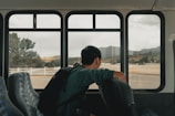 A person with a backpack sits alone on a bus, gazing out of the window at an open landscape with hills and scattered trees. The interior of the bus has empty seats with a patterned design.