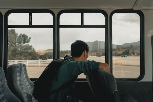 A person with a backpack sits alone on a bus, gazing out of the window at an open landscape with hills and scattered trees. The interior of the bus has empty seats with a patterned design.