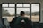A person with a backpack sits alone on a bus, gazing out of the window at an open landscape with hills and scattered trees. The interior of the bus has empty seats with a patterned design.
