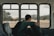 A person with a backpack sits alone on a bus, gazing out of the window at an open landscape with hills and scattered trees. The interior of the bus has empty seats with a patterned design.