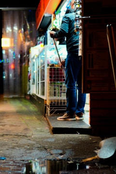 A person wearing casual clothing is sweeping the ground outside a small, illuminated shop with various items displayed in the windows. The surrounding area is wet, with a puddle reflecting the colorful lights of the shop. The scene takes place at night, with the area lit by streetlights and shop lights.