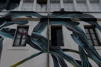 A building facade featuring large, stylized fish painted in dark shades with blue and green highlights. The fish's elongated shapes intertwine around the windows and walls. There are several windows on the building, some of which are open, and the overall atmosphere seems urban and artistic.