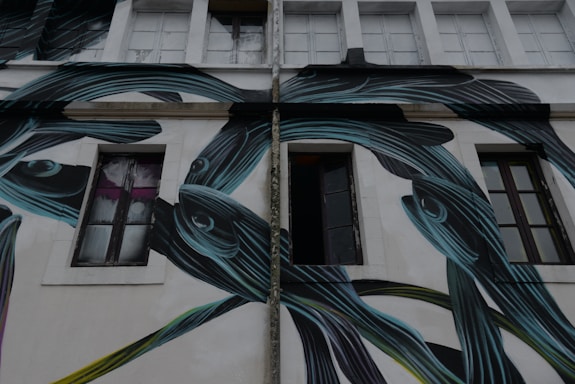 A building facade featuring large, stylized fish painted in dark shades with blue and green highlights. The fish's elongated shapes intertwine around the windows and walls. There are several windows on the building, some of which are open, and the overall atmosphere seems urban and artistic.
