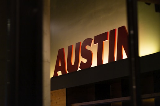 Large red letters spelling 'AUSTIN' are mounted on a dark surface with a subtle warm backlight creating a shadow effect on the wall. The setting appears to be indoors, with minimal ambient lighting.
