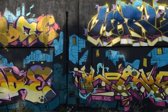 A vibrant graffiti mural on a wall, featuring bold and colorful lettering with abstract shapes. The backdrop includes blue urban skyline imagery with stylized buildings and windows, giving a sense of depth and contrast.