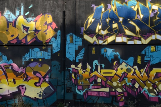 A vibrant graffiti mural on a wall, featuring bold and colorful lettering with abstract shapes. The backdrop includes blue urban skyline imagery with stylized buildings and windows, giving a sense of depth and contrast.