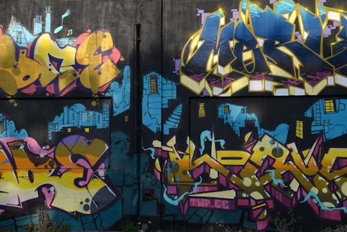 A vibrant graffiti mural on a wall, featuring bold and colorful lettering with abstract shapes. The backdrop includes blue urban skyline imagery with stylized buildings and windows, giving a sense of depth and contrast.
