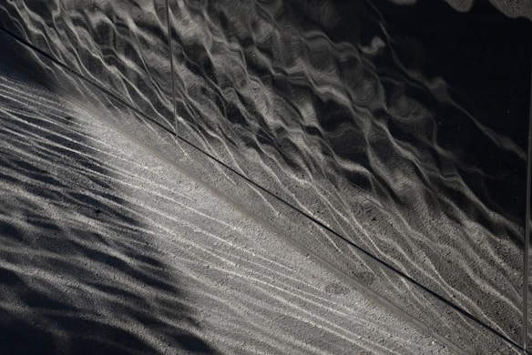 A textured surface with wavy, metallic reflections juxtaposed with a grainy, concrete-like texture. The lighting creates a contrast between shiny and matte areas, emphasizing the intricate patterns and lines.