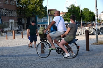A man and a woman are riding a bicycle together in an urban street setting. The woman is seated on the back, while the man pedals. They are casually dressed, suggesting a relaxed outing. Nearby, a pedestrian is walking, and there are trees and buildings in the background. A ferris wheel is visible in the distance.