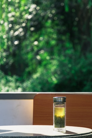 A glass bottle filled with tea and tea leaves sits on a round table, illuminated by sunlight. A lush, green, blurred background creates a peaceful outdoor setting. The bottle is transparent, with a metallic lid, allowing the golden tea to catch the light beautifully.