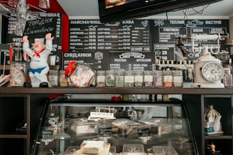 A café counter featuring a variety of items. On the top shelf, there is a ceramic figure of a chef, a scale, various glass jars containing herbs or spices, and other small décor items. Below, the display case contains various pastries and desserts. The background includes a blackboard menu with numerous items written in Cyrillic, listing coffee, breakfast, and other offerings.