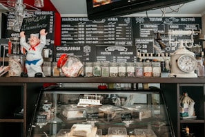 A café counter featuring a variety of items. On the top shelf, there is a ceramic figure of a chef, a scale, various glass jars containing herbs or spices, and other small décor items. Below, the display case contains various pastries and desserts. The background includes a blackboard menu with numerous items written in Cyrillic, listing coffee, breakfast, and other offerings.