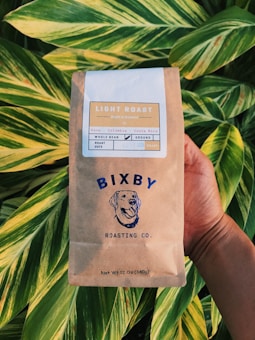 A hand holds a bag of Bixby Roasting Co. light roast coffee in front of large green leaves. The packaging is brown with blue and white designs, featuring a dog's face. Labels indicate the coffee is a mix from Kona, Colombia, and Costa Rica, and it is available as whole bean or ground.