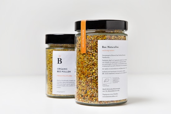 Two jars filled with colorful organic bee pollen, featuring black lids and white labels with text. The jar in the foreground has the label 'Bee Naturalles' with additional details and an orange tag. The second jar, partially visible in the background, is labeled 'Organic Bee Pollen'.