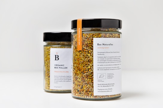Two jars filled with colorful organic bee pollen, featuring black lids and white labels with text. The jar in the foreground has the label 'Bee Naturalles' with additional details and an orange tag. The second jar, partially visible in the background, is labeled 'Organic Bee Pollen'.