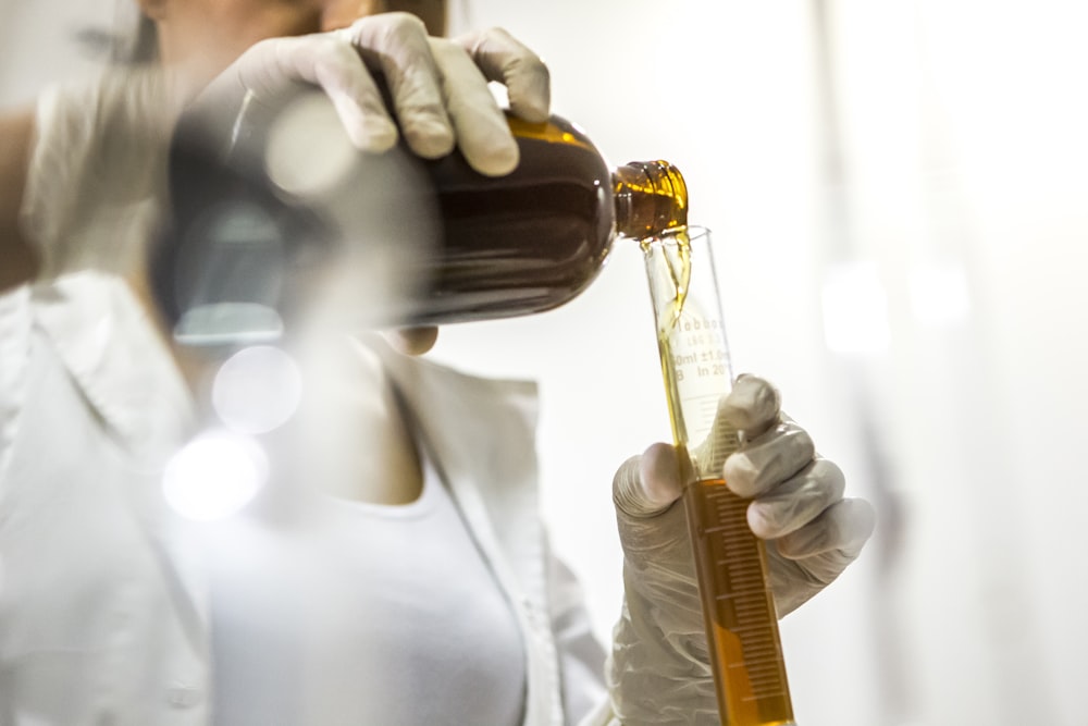 donna che indossa un camice da laboratorio bianco che tiene una bottiglia marrone e un tubo di vetro
