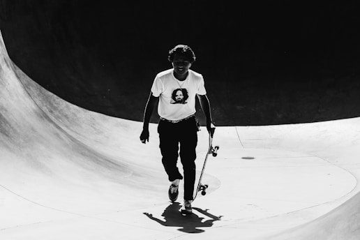 A person holding a skateboard walks through a skate park. The skateboarder is wearing a t-shirt with a printed design and the overall scene is captured in black and white.
