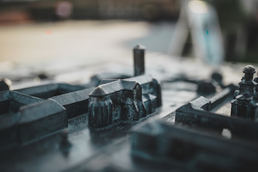 The image depicts a close-up view of a miniature model of a city, featuring small-scale buildings and structures made from a dark, metallic material. The focus is on the intricately detailed constructions, with some elements slightly out of focus, creating a sense of depth and realism.