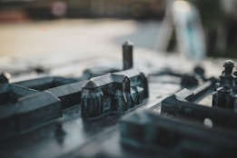 The image depicts a close-up view of a miniature model of a city, featuring small-scale buildings and structures made from a dark, metallic material. The focus is on the intricately detailed constructions, with some elements slightly out of focus, creating a sense of depth and realism.