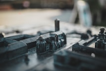 The image depicts a close-up view of a miniature model of a city, featuring small-scale buildings and structures made from a dark, metallic material. The focus is on the intricately detailed constructions, with some elements slightly out of focus, creating a sense of depth and realism.
