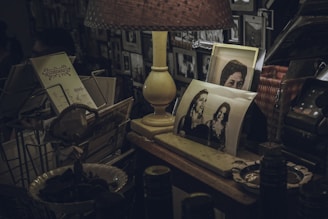 A dimly lit room features a table cluttered with nostalgic items. A vintage lamp emits a warm light, illuminating several black-and-white photographs, some portraying people smiling. A wire rack holds old postcards or letters. Various other objects including a retro telephone and decorative items surround the scene, suggesting a feeling of warmth and history.