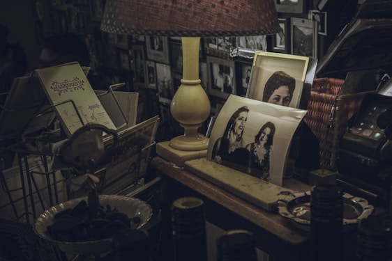 A dimly lit room features a table cluttered with nostalgic items. A vintage lamp emits a warm light, illuminating several black-and-white photographs, some portraying people smiling. A wire rack holds old postcards or letters. Various other objects including a retro telephone and decorative items surround the scene, suggesting a feeling of warmth and history.