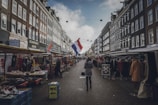 A bustling street market lined with stalls on either side, offering various goods and clothing. The scene is framed by traditional European buildings, with a prominent Dutch flag hanging above the street. A few people are walking through the market, browsing the items on display under the overcast sky.