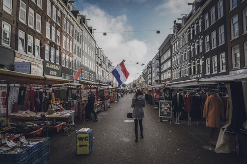 A bustling street market lined with stalls on either side, offering various goods and clothing. The scene is framed by traditional European buildings, with a prominent Dutch flag hanging above the street. A few people are walking through the market, browsing the items on display under the overcast sky.