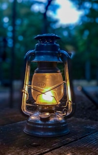 Close-up of a handcrafted camping lantern glowing warmly against a backdrop of pine trees.