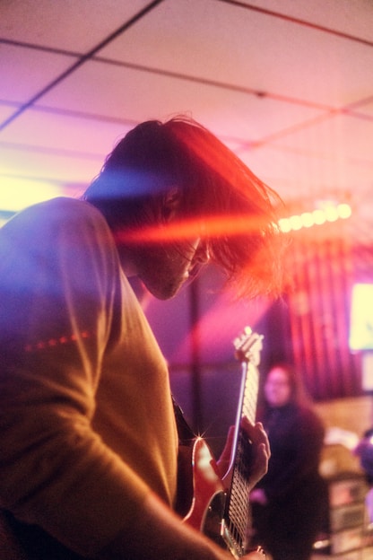 A musician with long hair is playing an electric guitar. The lighting creates colorful beams, including reds and blues, which add a dramatic effect to the scene. Another person is visible in the blurred background, suggesting a performance or concert setting.
