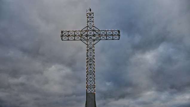 A large, metallic cross structure stands prominently against a backdrop of thick, gray clouds. Small lights line the edges of the cross, emitting a subtle glow. The atmosphere is moody and overcast, adding a dramatic ambiance to the scene.