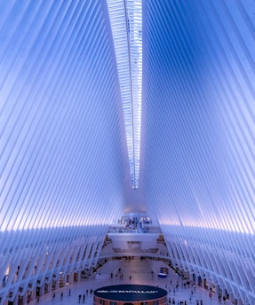 A vast, open interior space with a futuristic architectural design, featuring rows of white, rib-like structures that form a symmetrical and elongated ceiling. Natural light floods through a strip of windows at the top, casting a soft blue hue throughout the space. People are visible at various points, walking along the expansive floor and upper levels.