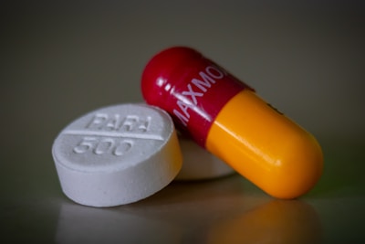 A close-up of two different types of medication. One object is a white round pill with 'PARA 500' written on it. The other is a red and yellow capsule with text inscribed on it.